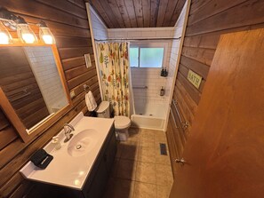 Ein Badezimmer mit Holzvertäfelung, einem Waschbecken mit Spiegel, einer Toilette, einer Badewanne und einem Duschvorhang mit Blumenmuster.