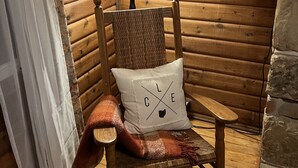 Interieur van een houten blokhut met een rieten stoel, een lamp en een kussen met 'CLE' en een zwart omtrek van een deelstaat.