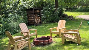 Houten Adirondack-stoelen staan rond een ronde vuurkuip van baksteen, in een weelderige tuin.