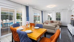 A modern living room with a wooden dining table set for four, a beige sofa, and a large window overlooking a swimming pool. - Villa Vista - Four Bedroom Villa, Sleeps 8 (Dalyan)