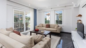 A modern living room with a sofa, a coffee table, and a TV. There are large windows with curtains, a balcony, and a view of houses. - Villa Vista - Four Bedroom Villa, Sleeps 8 (Dalyan)