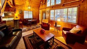 Interior acogedor de una cabaña con comedor, chimenea, sofá y un ventanal que ofrece vistas al exterior.