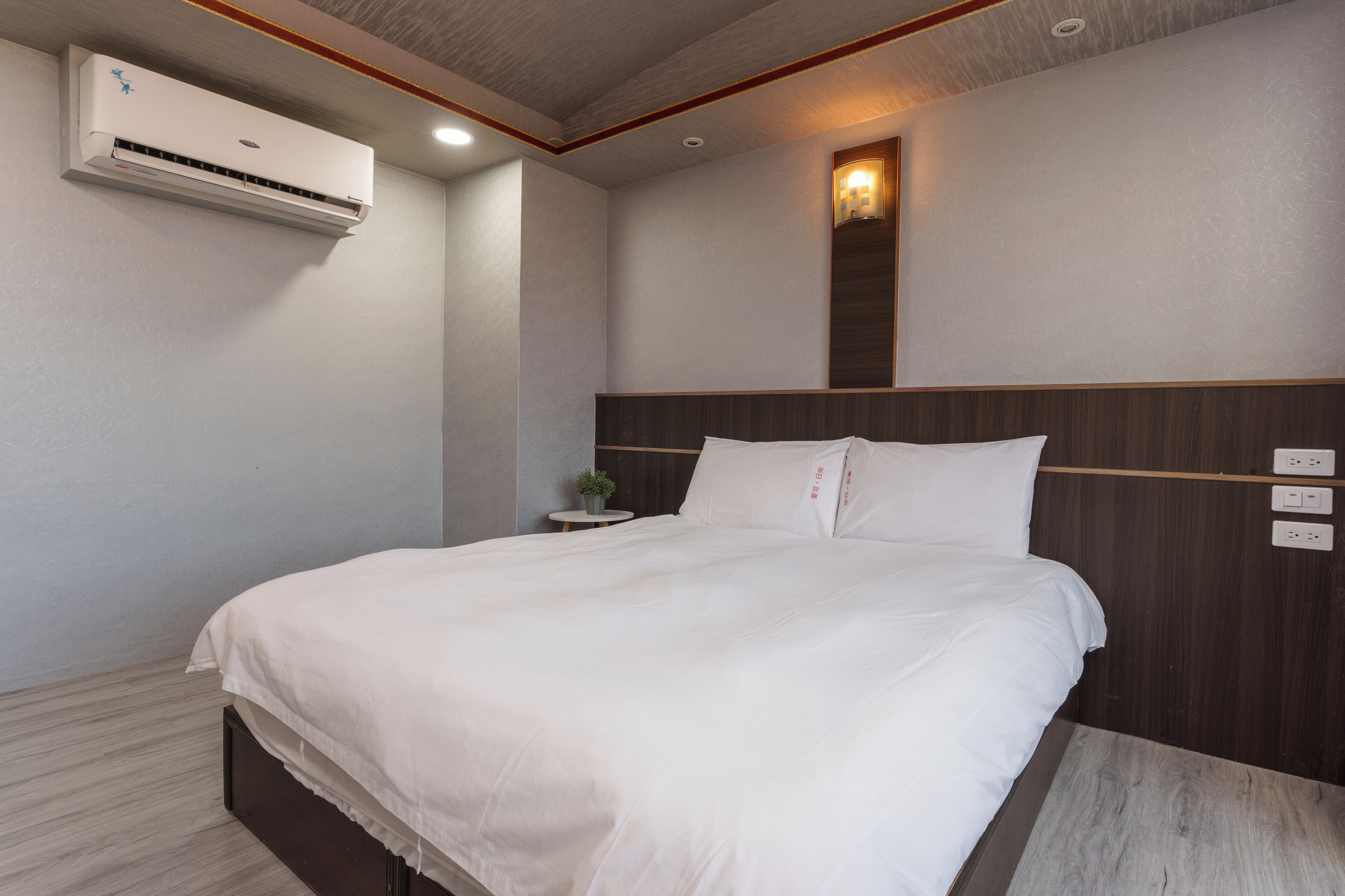 A modern hotel room with a large bed, an air conditioning unit, a small table with a plant, and a wall-mounted light fixture.