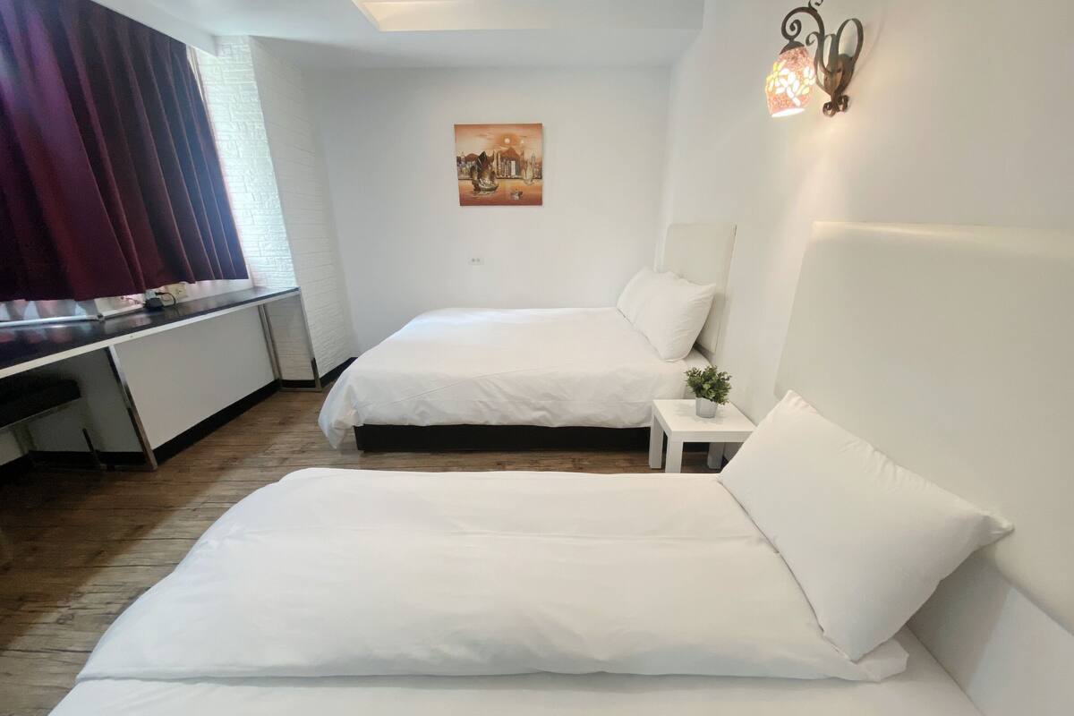 Ein Hotelzimmer mit zwei Betten, einem Schreibtisch, einem kleinen Tisch mit Pflanze und einem Bild an der Wand.