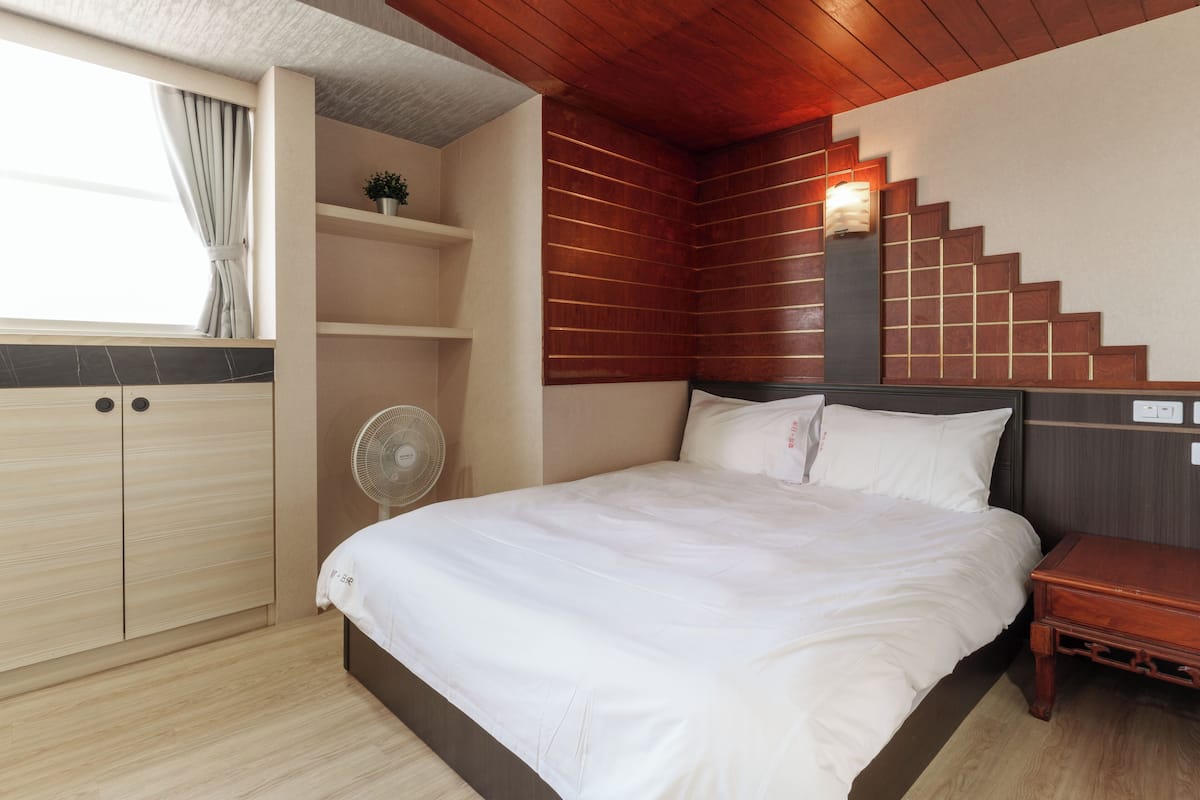 Ein Schlafzimmer mit einem Bett, einem Holzkopfende, einem Nachttisch, einem Ventilator, einem Regal mit einer Pflanze und einer Treppe mit einer Leuchte.