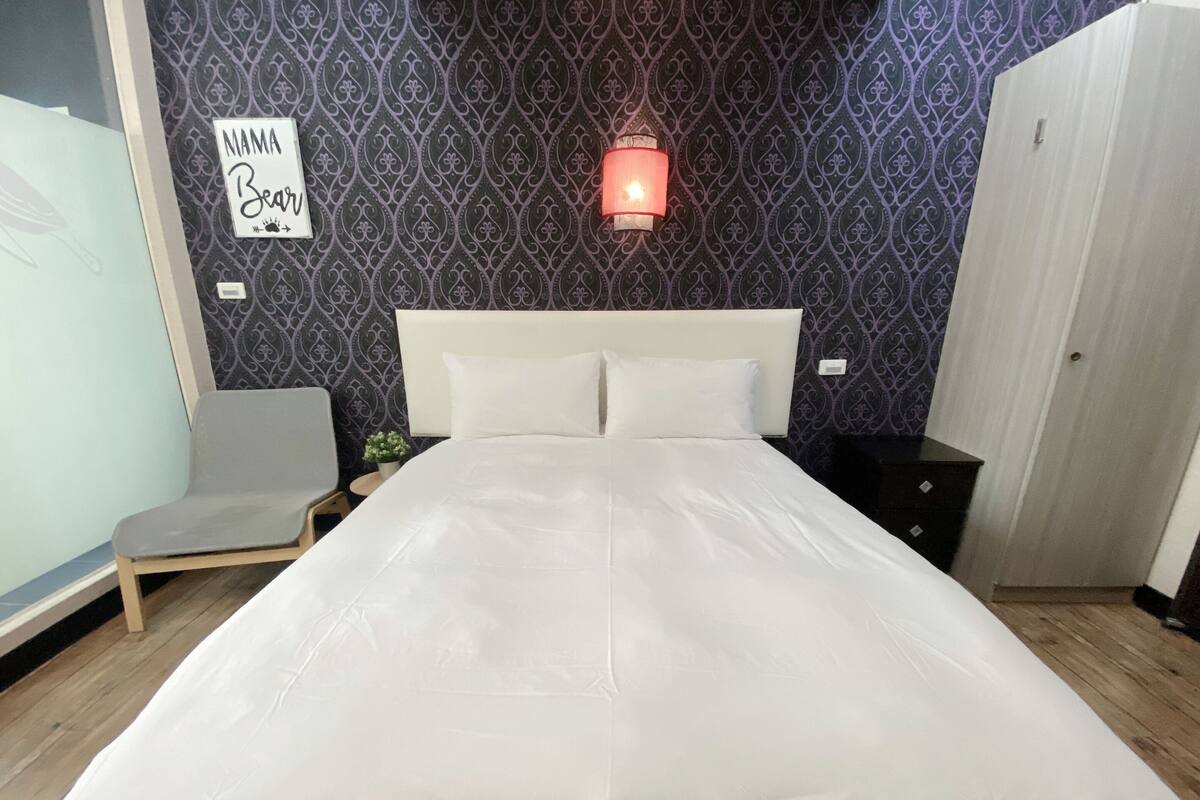 Ein ordentlich bezogenes Bett mit weißen Leinen, ein Nachttisch mit Lampe und ein Sessel in einem Zimmer mit gemustertem Wandpapier und einem Schild, das "NAMA Bear" zeigt.