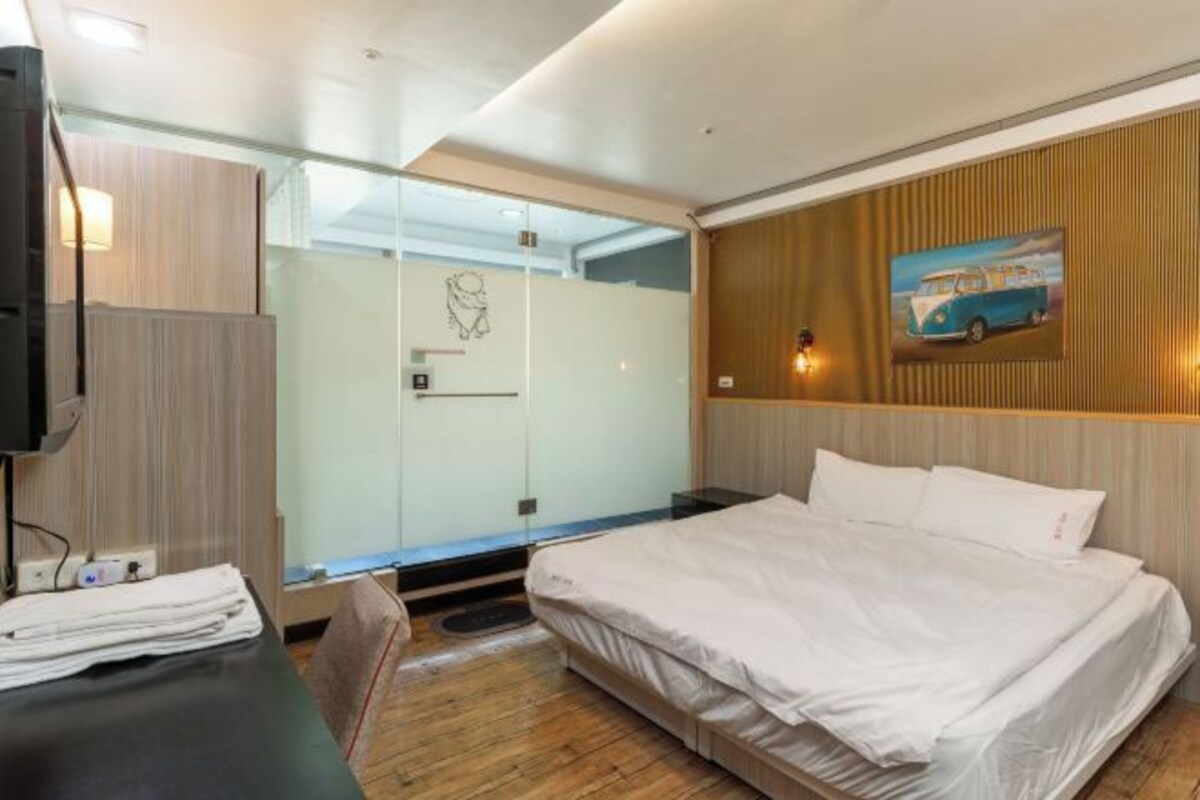Una habitación de hotel moderna con cama, escritorio con silla, cabina de ducha de vidrio y televisor montado en la pared.