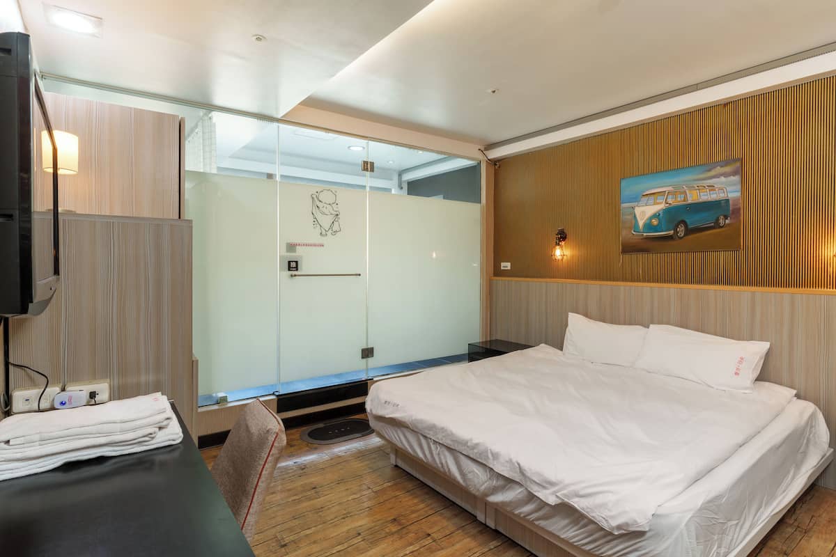 Ein modernes Hotelzimmer mit einem Bett, einem Schreibtisch, einem Sessel, einer Glasdusche und einem an der Wand befestigten Fernseher.