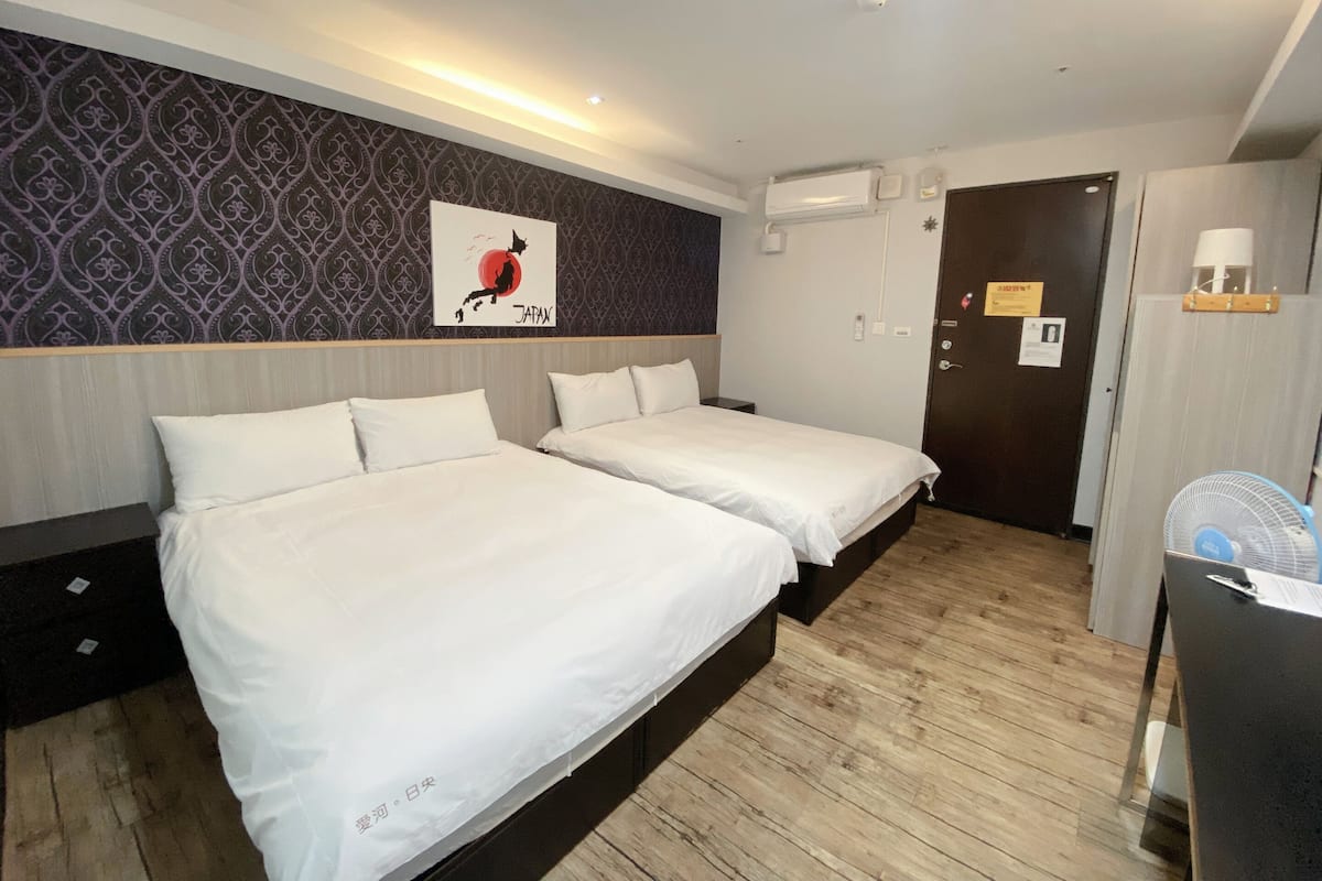 Ein Hotelzimmer mit zwei Betten, einer Wand-Klimaanlage, einer Tür mit einem Schild und einem kleinen Schreibtisch mit Ventilator.