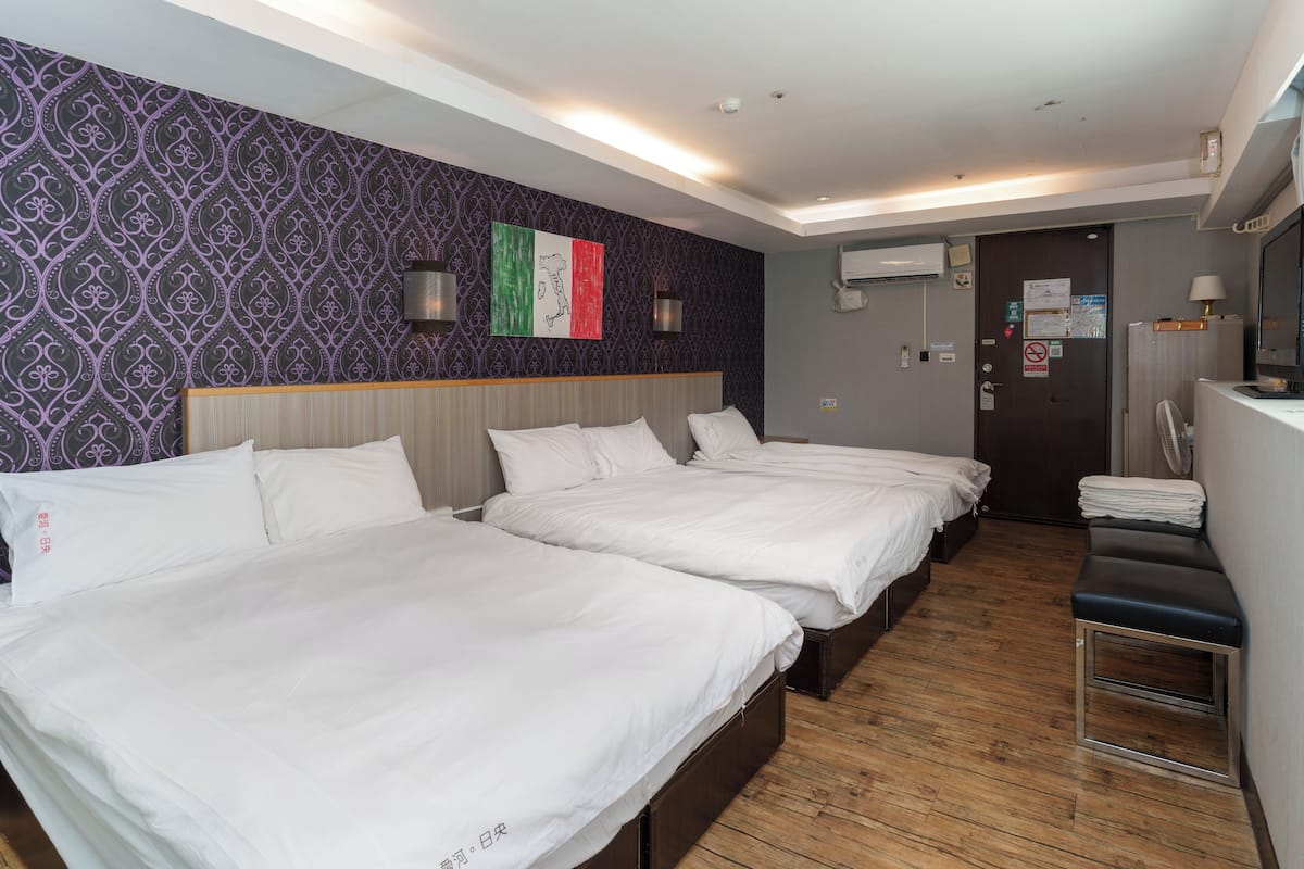 Ein Hotelzimmer mit drei Einzelbetten, Holzboden, einer Wand mit violettem Muster und einer Tür mit einem Schild.