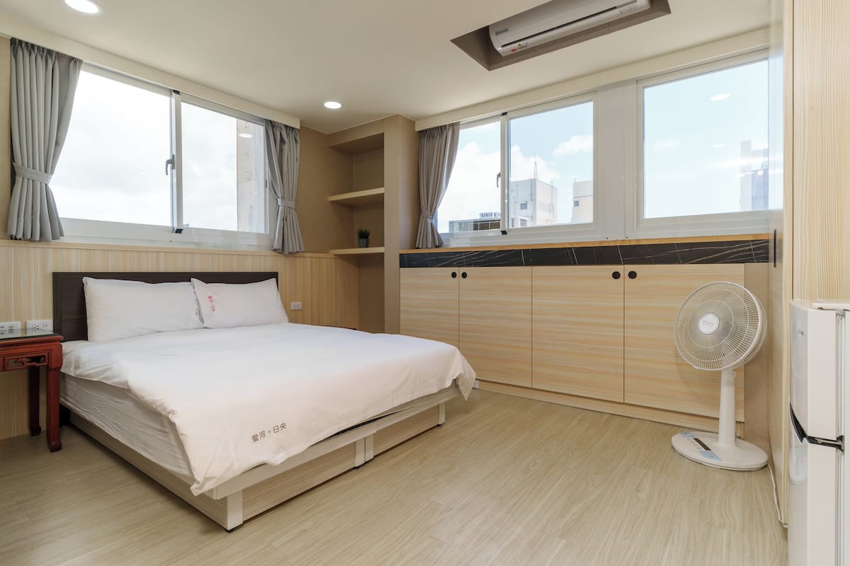 Ein Hotelzimmer mit Bett, Nachttisch, Ventilator und einem Fenster mit Vorhängen.