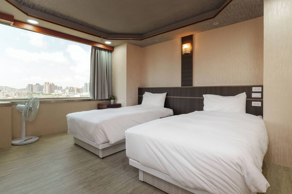 Ein Hotelzimmer mit zwei Betten, einem Standventilator, einem Nachttisch und einem Fenster mit Blick auf die Stadt.
