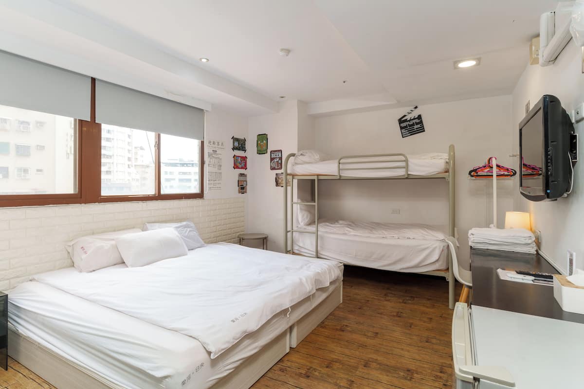 Ein Schlafsaalzimmer mit Etagenbetten, einem Schreibtisch, einem Stuhl, einem Fernseher und einem Fenster mit Blick auf Gebäude.