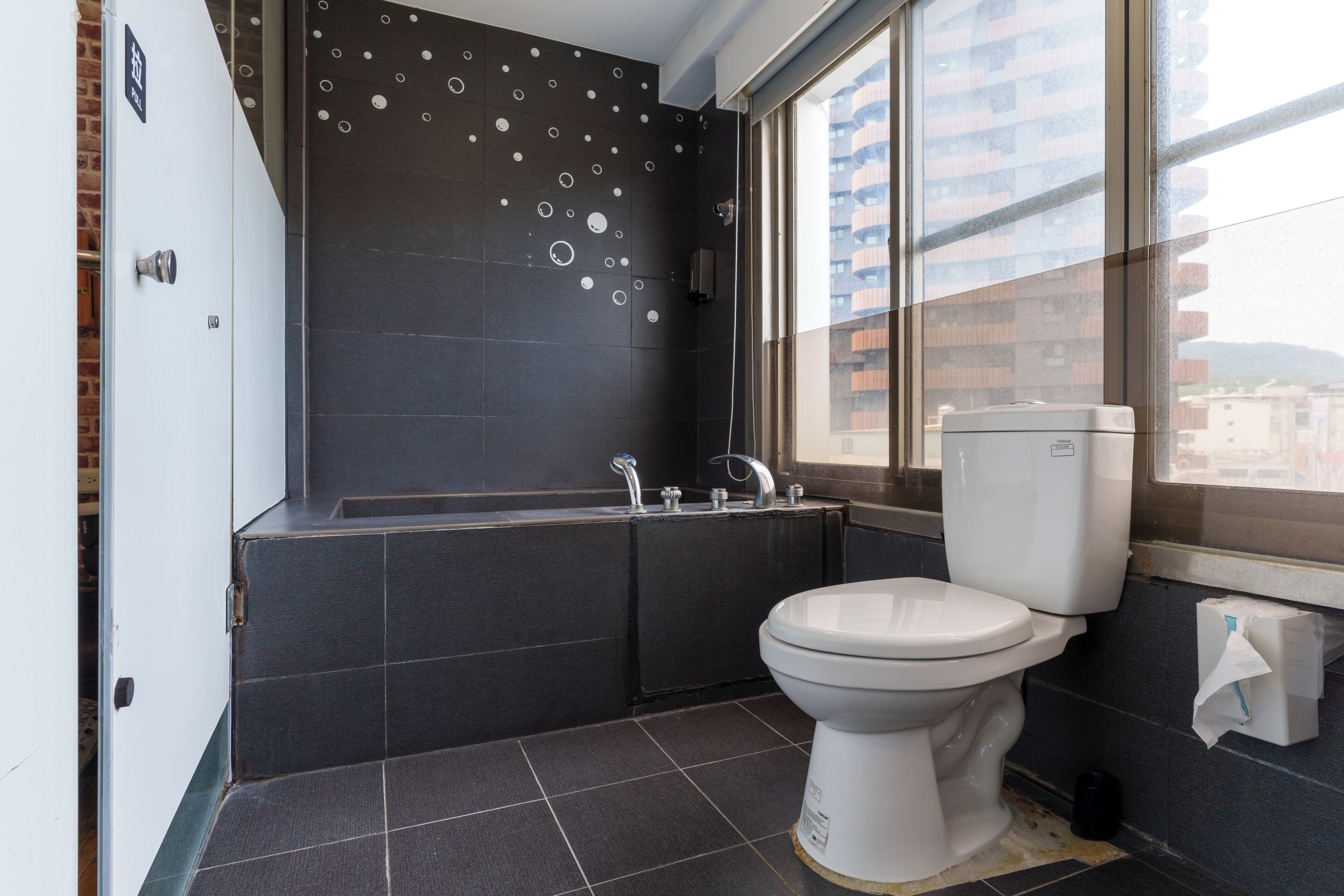 A modern bathroom with a bathtub, toilet, and a large window offering a view of buildings.
