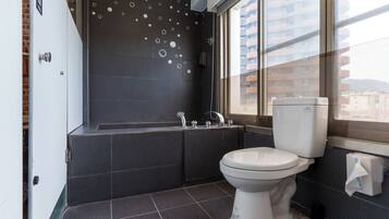 A modern bathroom with a bathtub, toilet, and a large window offering a view of buildings.