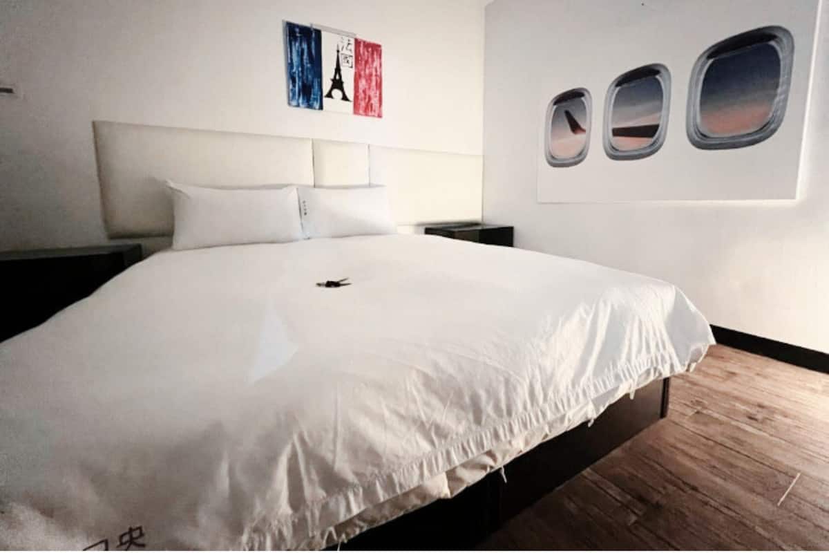 Ein Hotelzimmer mit einem großen Bett, einem Kopfteil, einem Nachttisch und einer Wand mit drei Fenstern.