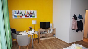 A hotel room with a yellow wall, a painting of paint bottles, a TV, a coffee machine, a desk with a lamp, and a bed with towels. - Posthus Apartment 103, close to the beach and central (Borkum)
