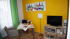 A room with a dining table, chairs, a TV, a coffee machine, and a shelf with electronic devices. - Posthus Apartment 103, close to the beach and central (Borkum)