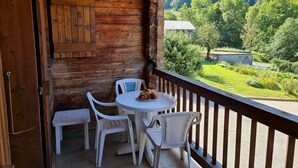 A wooden balcony with a table and chairs, overlooking a lush green valley and forested mountains. - Chalet Genépi - Apartment 1 (Le Grand-Bornand)