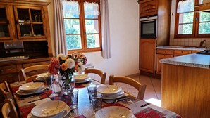 A dining area with a table set for six, wooden cabinets, a kitchen with an oven, and a window with curtains. - Chalet Genépi - Apartment 1 (Le Grand-Bornand)