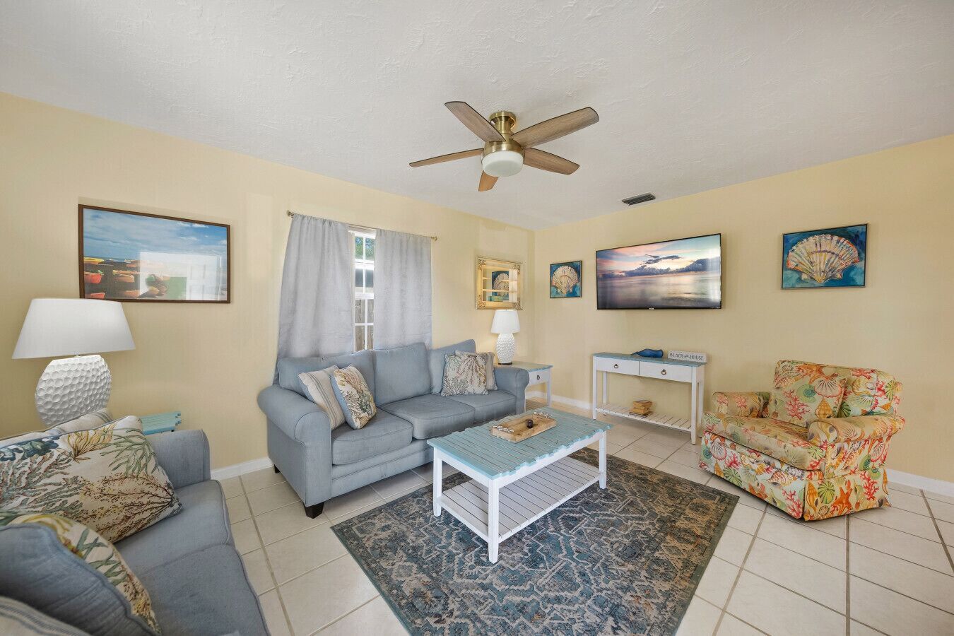 A living room with a grey sofa, a patterned armchair, a coffee table, a ceiling fan, and artwork on the walls.