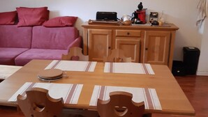 Een gezellig eetgedeelte met een houten tafel en stoelen, een rode bank, een keukenhoek met een klok, en een lijst met een afbeelding aan de muur.