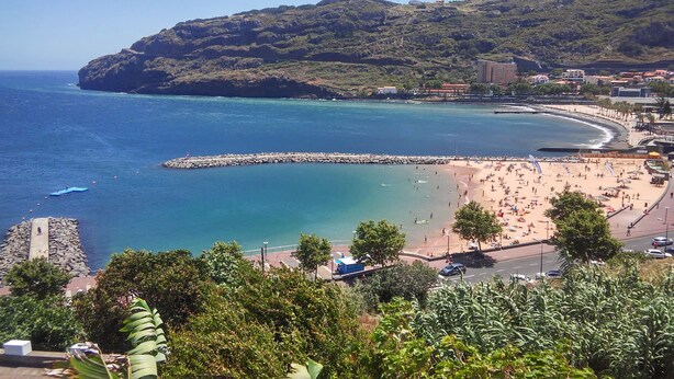 A coastal beach with a rocky breakwater, a sandy beach, and a hilly backdrop. - Sunny Beach Machico Apartment (Machico)