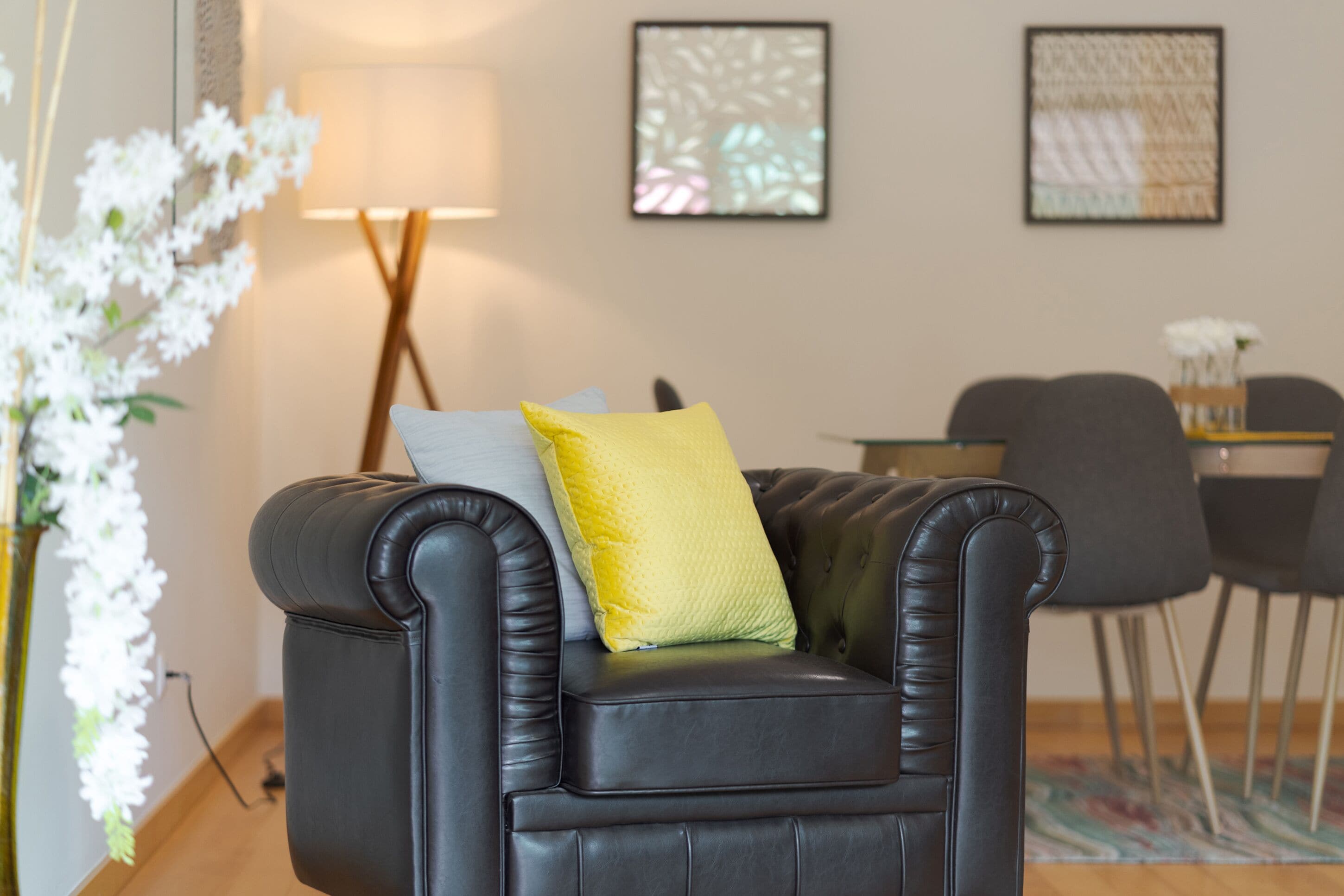 Een zwarte leren bank met een geel kussen en een grijs kussen, een lamp en twee ingelijste kunstwerken aan de muur.