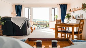 A cozy room with a bed, a dining table with chairs, a coffee table with cups, and a balcony with a view. - Spacious sea-view apt. w/ hot tub & sauna overlooking St Ives Bay (Hayle)