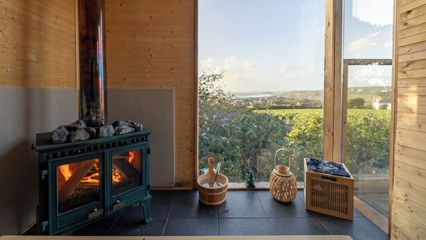 A cozy room with a wood-burning stove, a large window with a view of greenery, and a small table with a basket and a candle holder. - Spacious sea-view apt. w/ hot tub & sauna overlooking St Ives Bay (Hayle)
