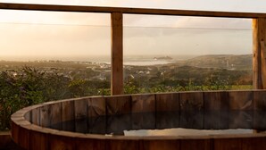 A wooden hot tub with a view of a coastal landscape, including hills and a lighthouse. - Spacious sea-view apt. w/ hot tub & sauna overlooking St Ives Bay (Hayle)