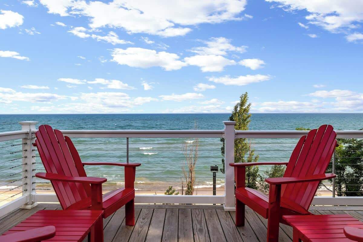 A wooden deck with two red Adirondack chairs facing a view of the ocean.