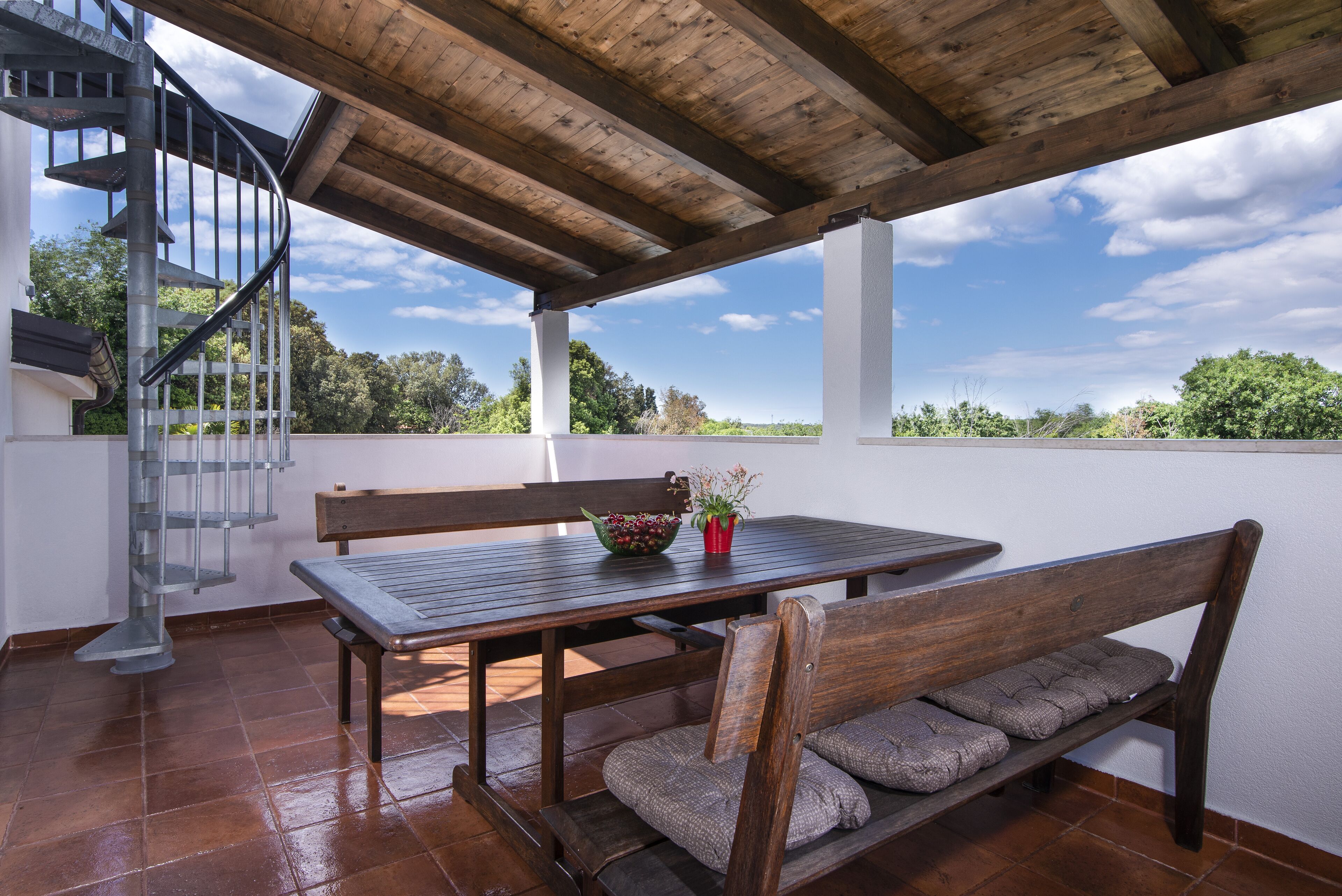 A covered patio with a wooden table and benches, a spiral staircase, and a view of the surrounding landscape.