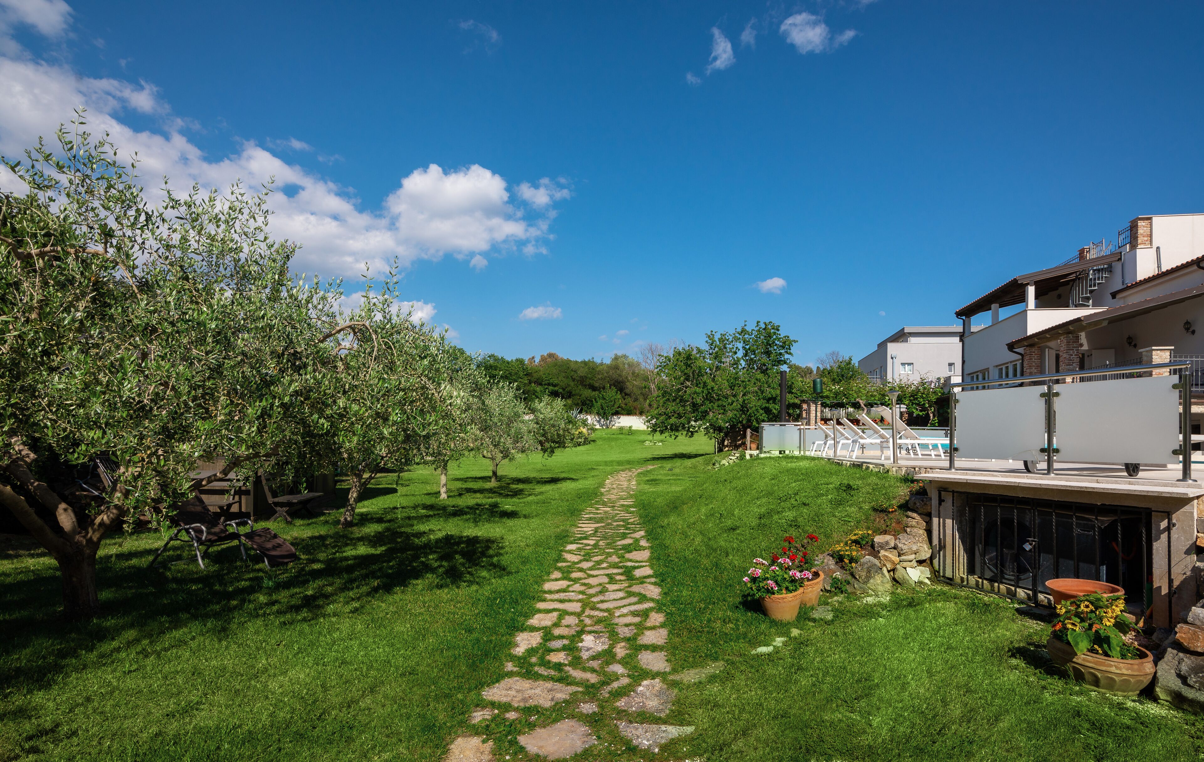 Polku, joka kulkee hyvin hoidetun puutarhan läpi ja johtaa uima-allasalueelle sekä rakennukseen, jossa on parveke.