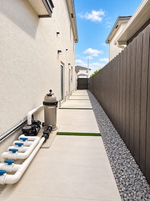 A narrow pathway flanked by a building and a wooden fence, with a water filtration system and a trash bin along the path. - Private & Comfy Efficiency (Miami)