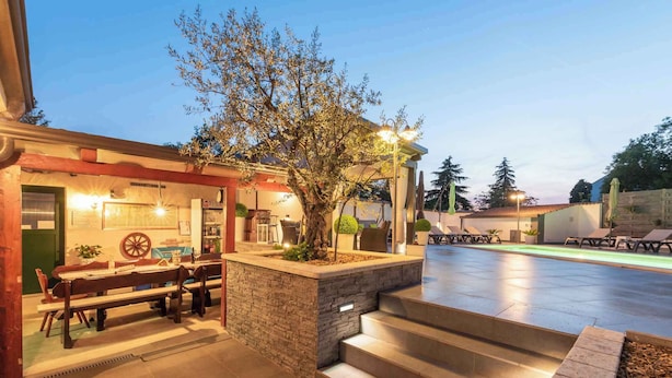 A well-lit outdoor area with a dining table and chairs, a stone planter with a tree, and a pool area with lounge chairs. - Holiday apartment with large garden and pool (Poreč)