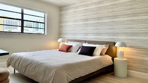 Ein modernes Schlafzimmer mit einem großen Bett, zwei Nachttischlampen und Blick auf die Stadt durch ein Fenster.