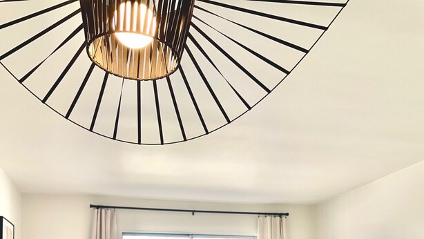 Ein modernes Wohnzimmer mit einer Couch, einem Esstisch und einem Fernseher. Es gibt Fenster mit Vorhängen, eine Deckenleuchte und einen Blick auf Grünflächen im Freien.