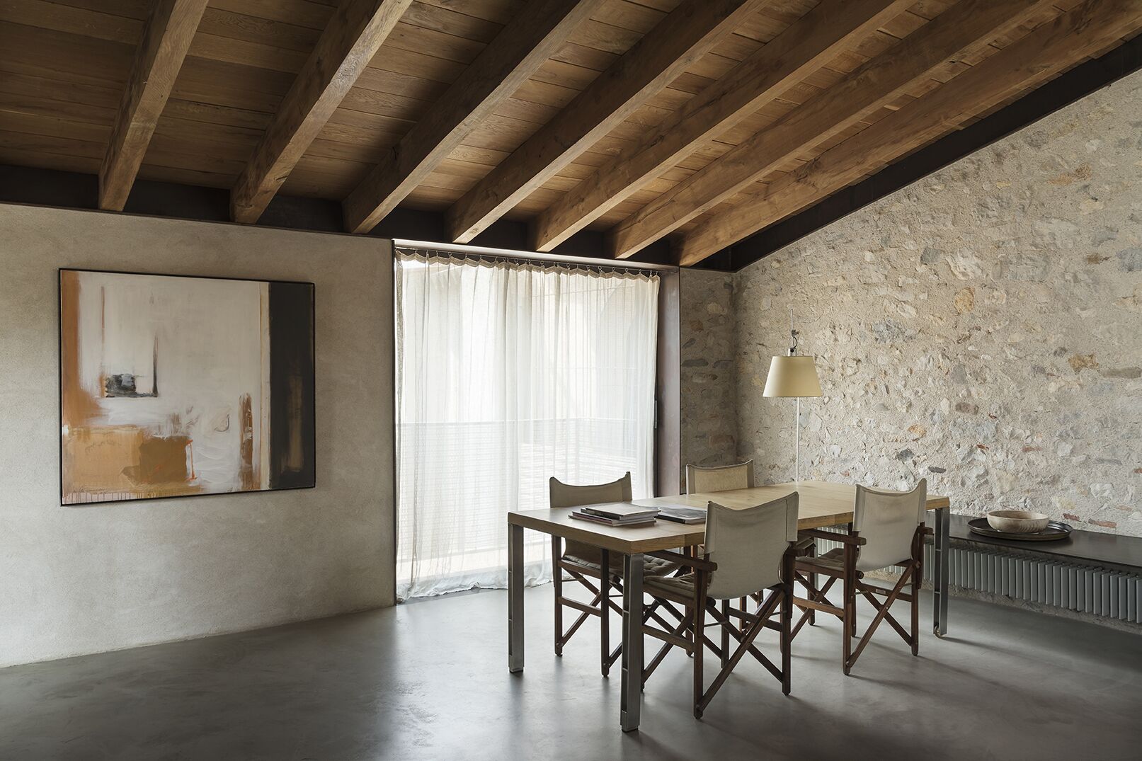 A dining area with a wooden table and chairs, a painting on the wall, a window with sheer curtains, and a stone wall.