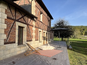 A wooden deck with a lounge chair and umbrella, a brick building with timber framing, and a grassy yard with trees.