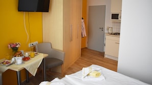 Ein Hotelzimmer mit Bett, Schreibtisch mit Frühstücksangeboten, Wandfernseher und einer Küchenzeile.