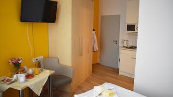 Ein Hotelzimmer mit Bett, Schreibtisch mit Frühstücksangeboten, Wandfernseher und einer Küchenzeile.