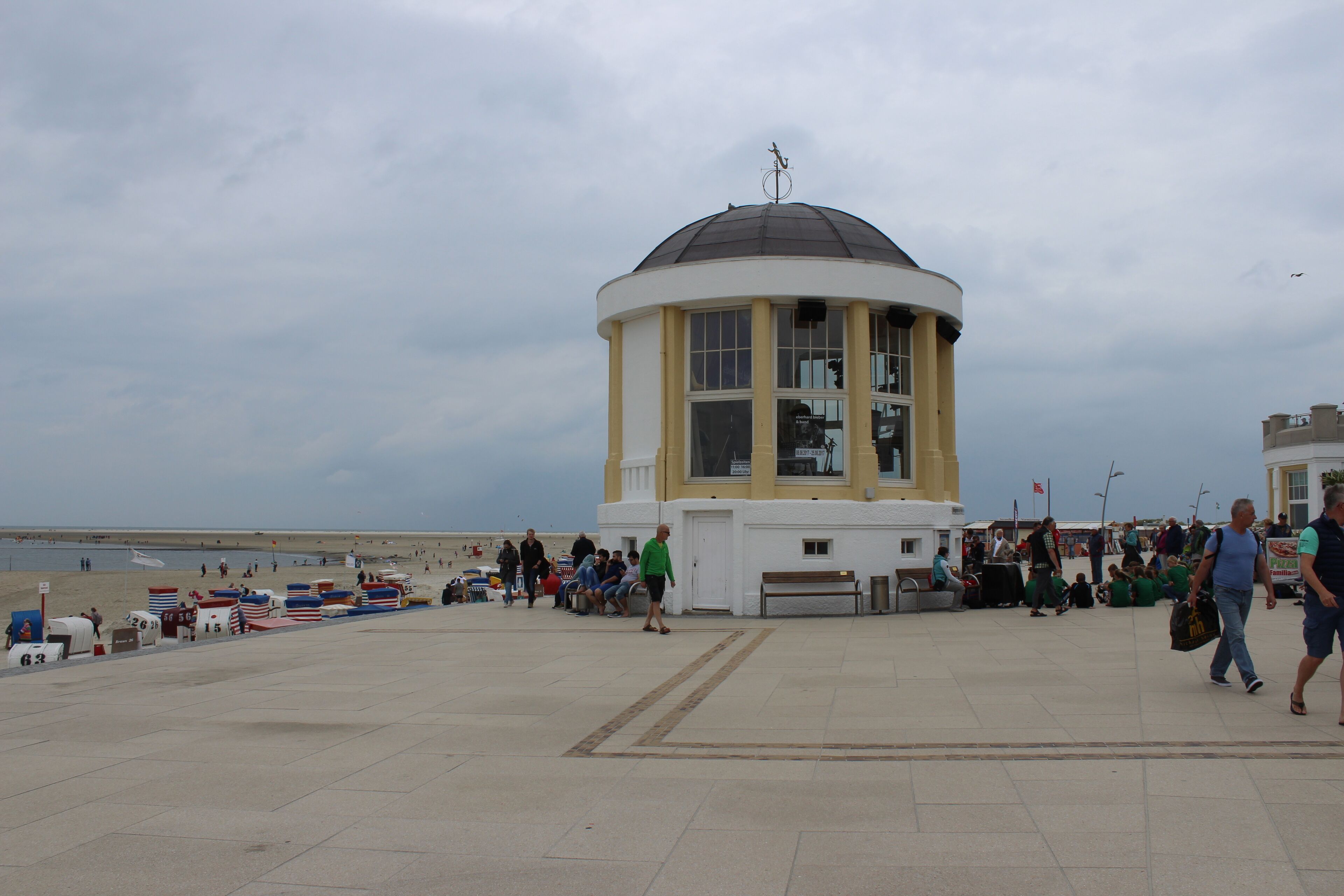 Eine Küstenpromenade mit einem markanten weiß-gelben Turm, Bänken und einem Strand im Hintergrund.