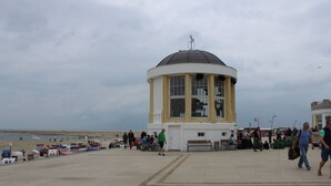 Eine Küstenpromenade mit einem markanten weiß-gelben Turm, Bänken und einem Strand im Hintergrund.