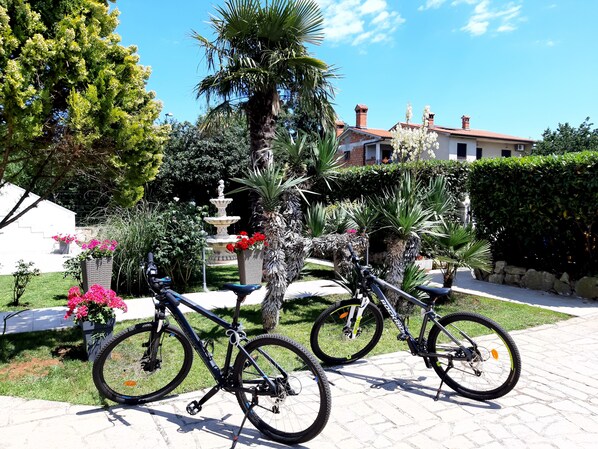 Deux vélos sont garés dans un jardin, avec une fontaine et un bâtiment en arrière-plan.