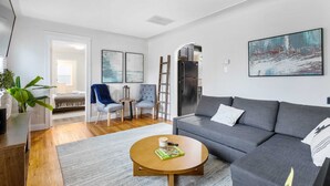 A modern living room with a grey sofa, a wooden coffee table, and a blue chair. There is a ladder leaning against the wall, a refrigerator, and a door leading to another room. - Stylish, Cute & Homey Livonia Getaway (Livonia)