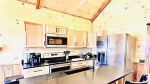 A kitchen with wooden cabinets, a central island, bar stools, and a microwave. - LUXURY COUPLES CABIN! Caveside Cabins (Creola)