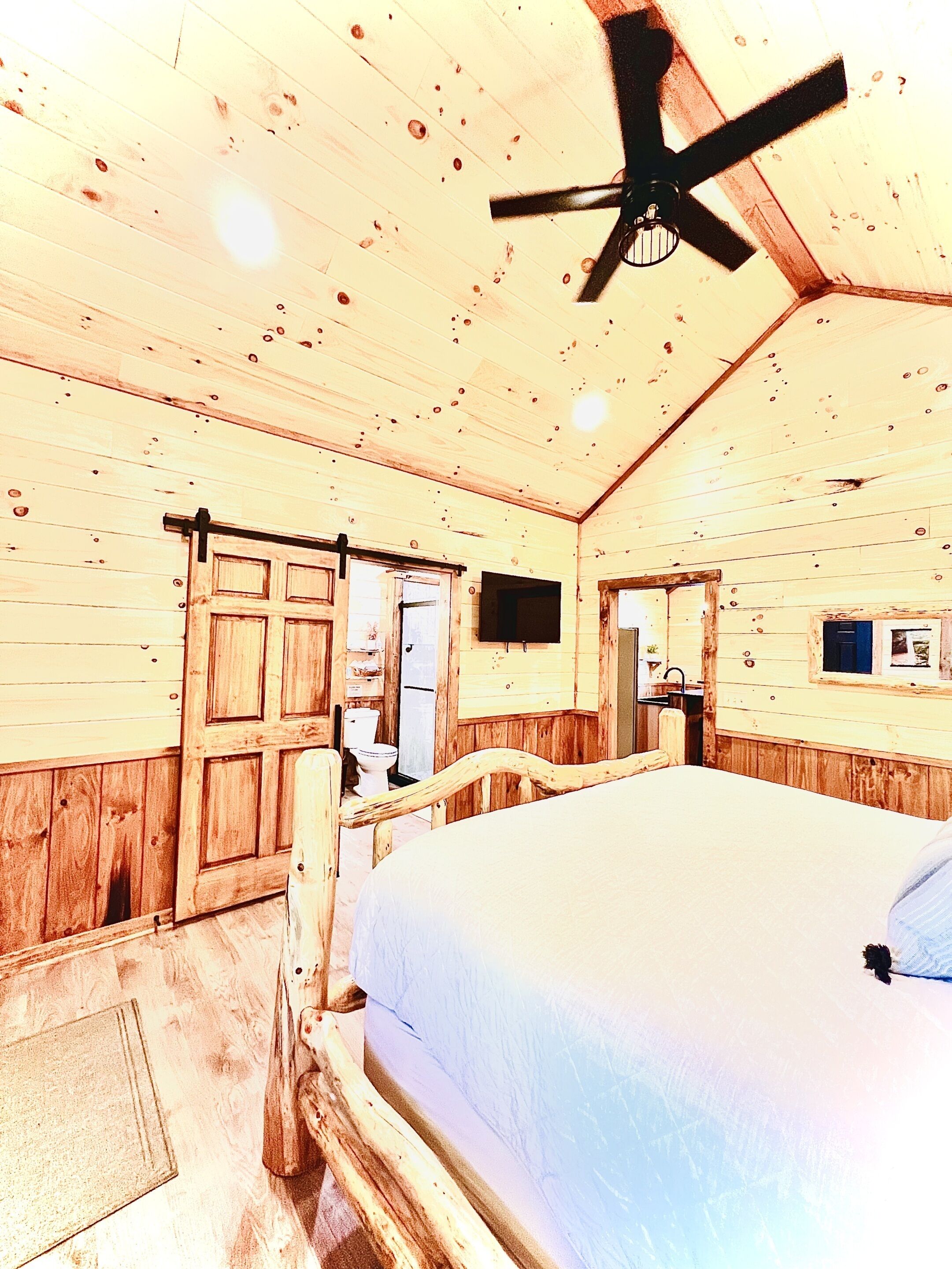 A wooden cabin bedroom with a bed, a ceiling fan, a wooden door, and a small window.