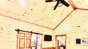 A wooden cabin bedroom with a bed, a ceiling fan, a wooden door, and a small window. - LUXURY COUPLES CABIN! Caveside Cabins (Creola)