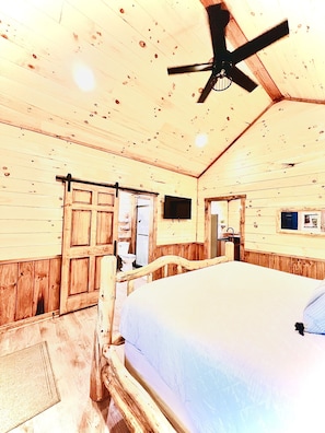 A wooden cabin bedroom with a bed, a ceiling fan, a wooden door, and a small window. - LUXURY COUPLES CABIN! Caveside Cabins (Creola)